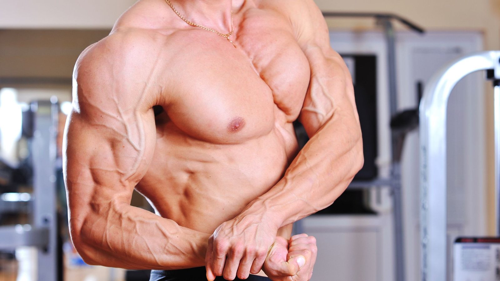 Muscular shirtless man flexing his bicep in a gym, veins prominent and a gold chain around his neck.